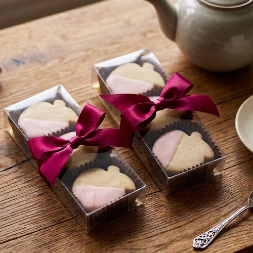 Two boxes of shortbread cookies with pink ribbons on a wooden table with a teapot and cup.