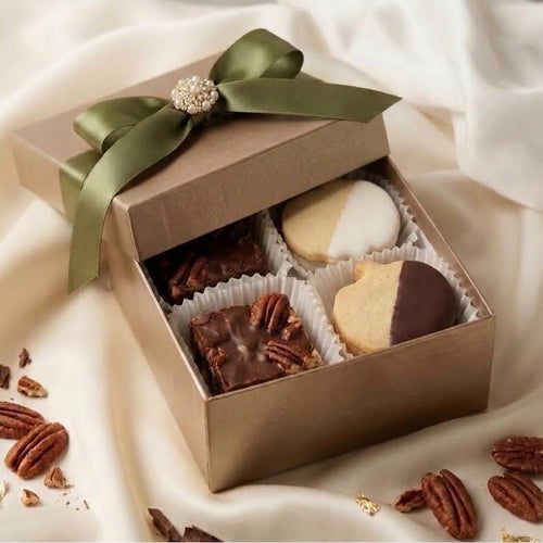 An open square brown gift box tied with an olive green satin ribbon and jeweled bow, filled with assorted pumpkin cut shortbread and pecan squares, set on draped cream fabric with scattered nuts around it.