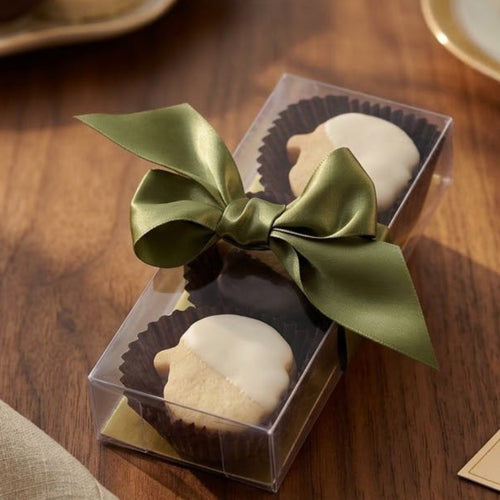 A clear plastic gift box containing 6 pumpkin shaped shortbread dipped in white and dark chocolate with an olive green satin bow, set on a wooden table with a beige fabric napkin, pecans, chocolate, and a handwritten gift tag.