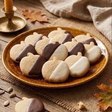 Pumpkin-shaped cookies, some plain and some half-chocolate and half-white icing, arranged on a ceramic plate on a rustic wooden table with autumn leaves, pumpkin seeds, a candle, and a beige cloth.